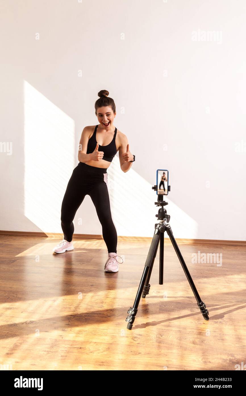 Fitness vlogger recording tutorials for sporty class, showing thumbs up to phone camera on tripod, wearing black sports top and tights. Full length studio shot illuminated by sunlight from window. Stock Photo