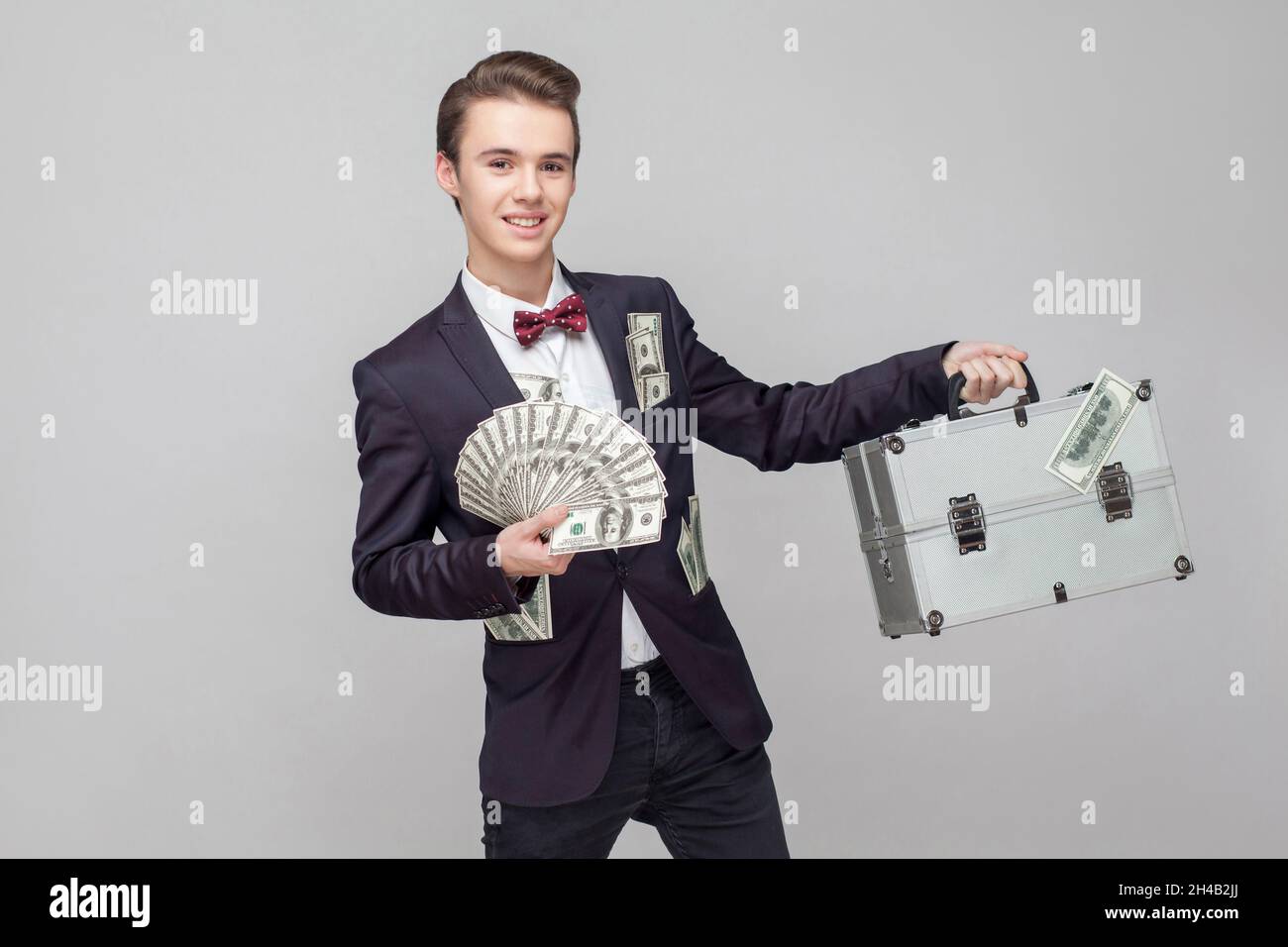 Portrait of cheerful excited millionaire, businessman with stylish hairdo in black suit with many dollars in pocket showing suitcase and bunch of money. indoor studio shot isolated on gray background Stock Photo