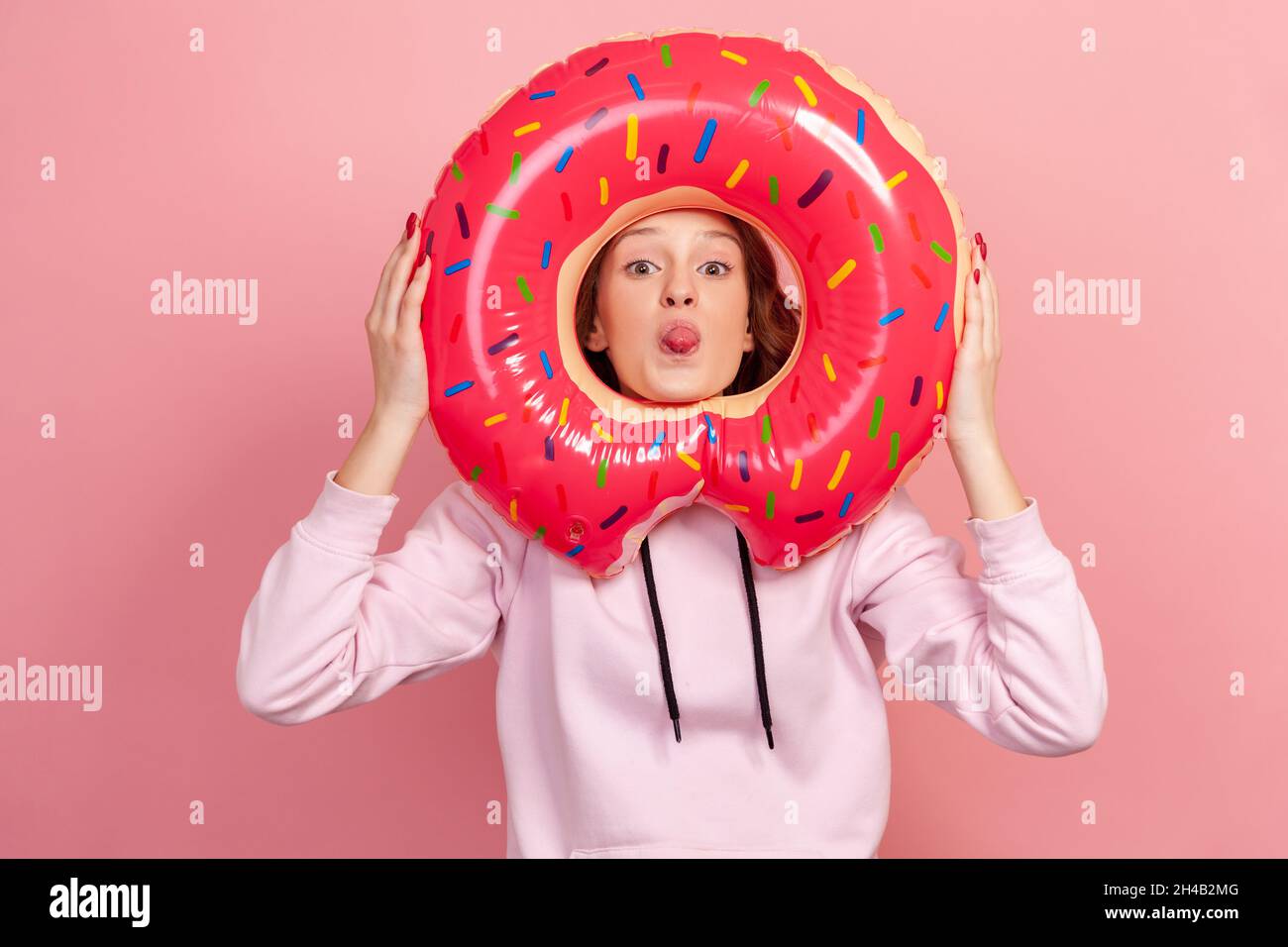 Portrait of funny curly haired teenage girl in hoodie showing tongue through pink donut rubber ring holding in hands, fooling around, rest. Indoor studio shot isolated on pink background Stock Photo