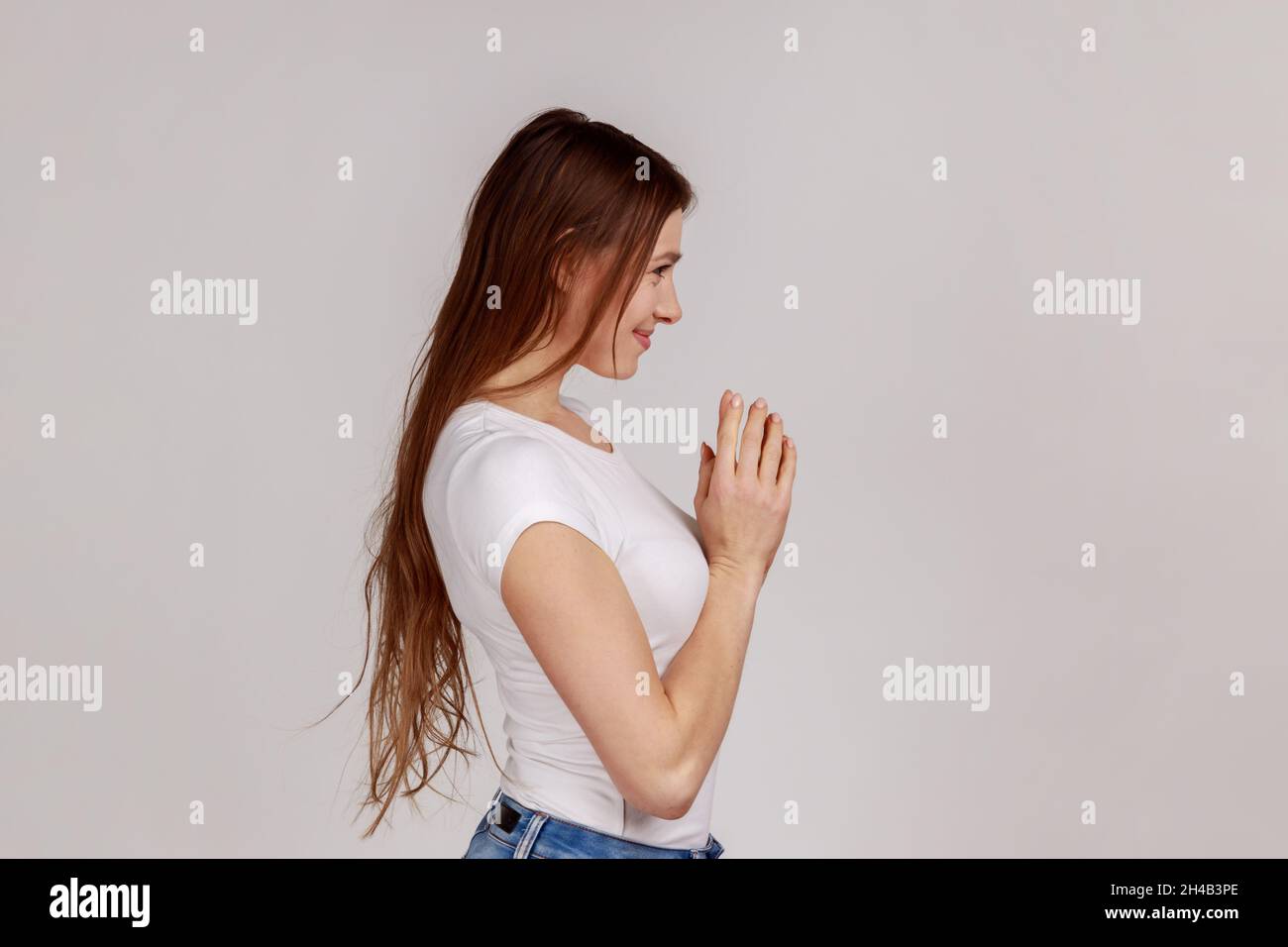 Side view portrait of scheming devious woman clasping hands and thinking over tricky plan, having sly cunning idea to prank, wearing white T-shirt. Indoor studio shot isolated on gray background. Stock Photo
