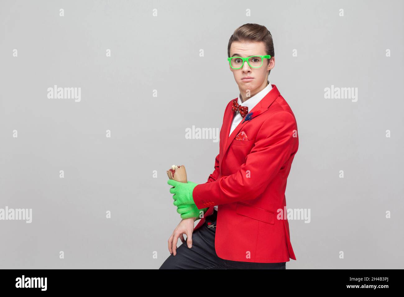 Portrait of stylish bizarre gentleman with eyeglasses in red tuxedo and green gloves holding zombie hand crawling over his leg, looking surprised at camera. studio shot isolated on gray background Stock Photo