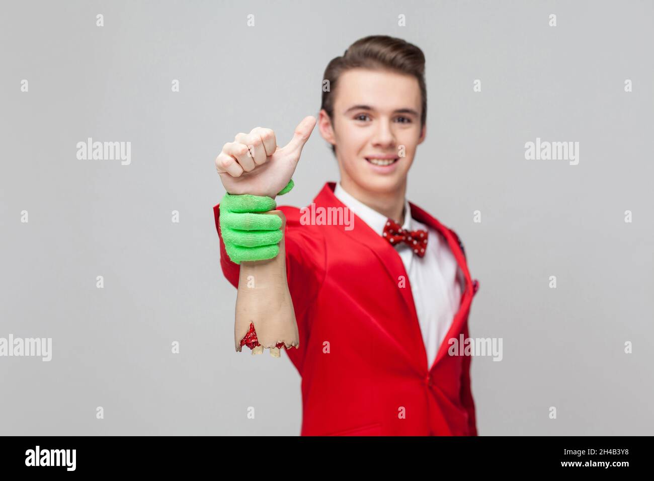 Portrait of cheerful gentleman with stylish hairdo in red tuxedo and bow tie smiling at camera and holding zombie hand showing thumbs up, like gesture. indoor studio shot isolated on gray background Stock Photo