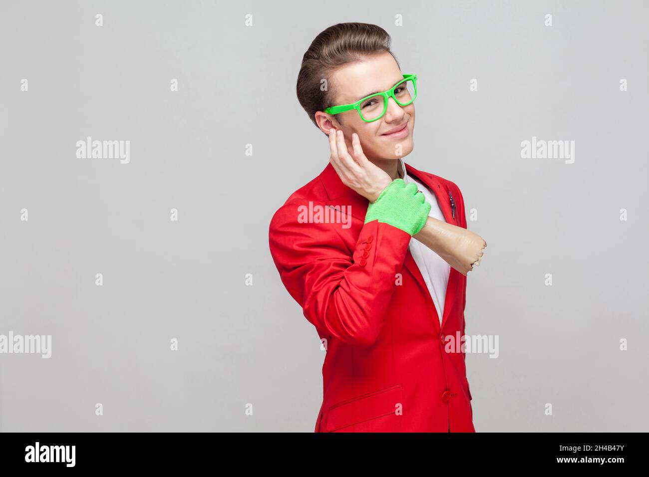 Portrait of positive funny gentleman with eyeglasses in red tuxedo and green gloves holding zombie hand, stroking his cheek or pretending to make phone call. studio shot isolated on gray background Stock Photo