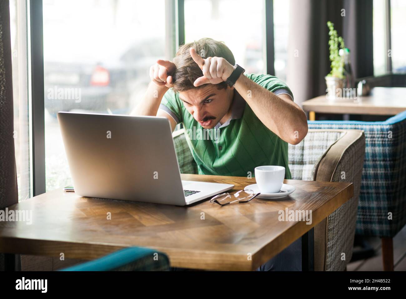 Portrait of angry businessman wearing green T-shirt at work, looking at laptop monitor, showing bull horn gesture, threatening to attack. Indoor shot near big window, cafe background. Stock Photo