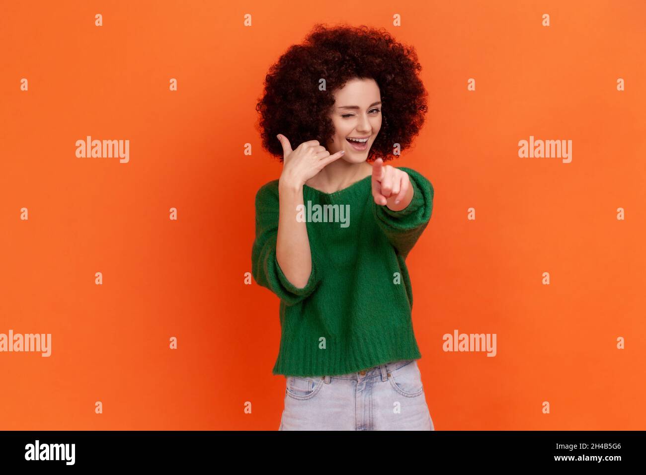 Hey you, call me. Woman with Afro hairstyle standing with telephone hand gesture and smiling to camera, flirting offering to contact by phone. Indoor studio shot isolated on orange background. Stock Photo