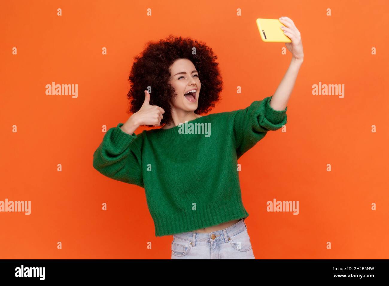 Excited happy woman with Afro hairstyle wearing green casual style sweater has livestream with followers, showing thumb up to camera. Indoor studio shot isolated on orange background. Stock Photo