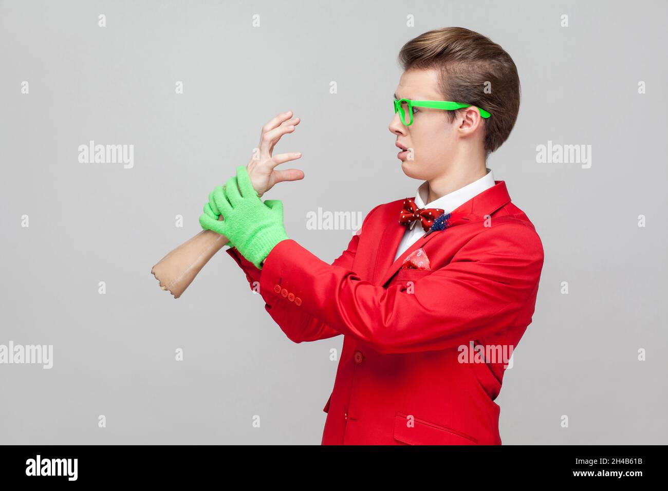 Portrait of bizarre funny gentleman with eyeglasses in red tuxedo and green gloves looking scared frightened and holding zombie hand trying to catch him. studio shot isolated on gray background Stock Photo