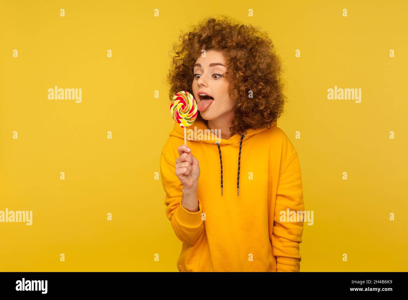 Attractive woman sticking out tongue licking lollipop, tasting sweet round candy, enjoying delicious flavor dessert, wearing casual style hoodie. Indoor studio shot isolated on yellow background. Stock Photo