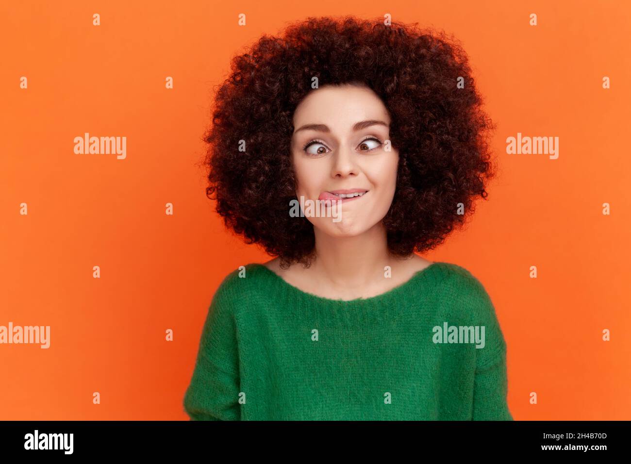 Foolish woman with Afro hairstyle wearing green casual style sweater standing with crossed eyes and tongue out, childish behavior. Indoor studio shot isolated on orange background. Stock Photo