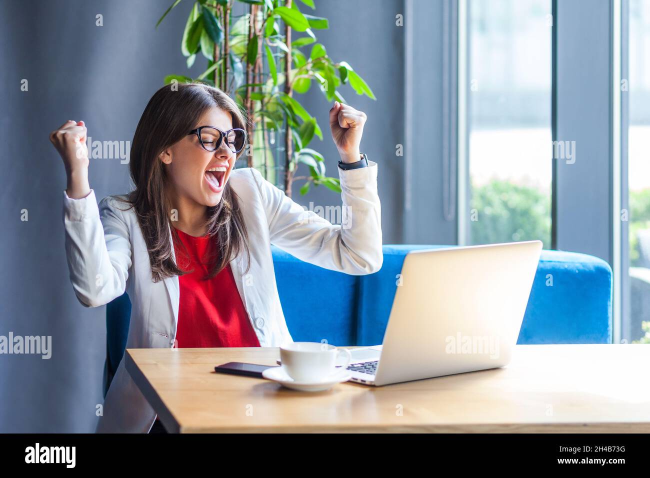 Extremely happy woman in glasses raising hands and screaming for joy, celebrating successful business online, rejoicing victory while working on laptop. Indoor shot, cafe or office background. Stock Photo