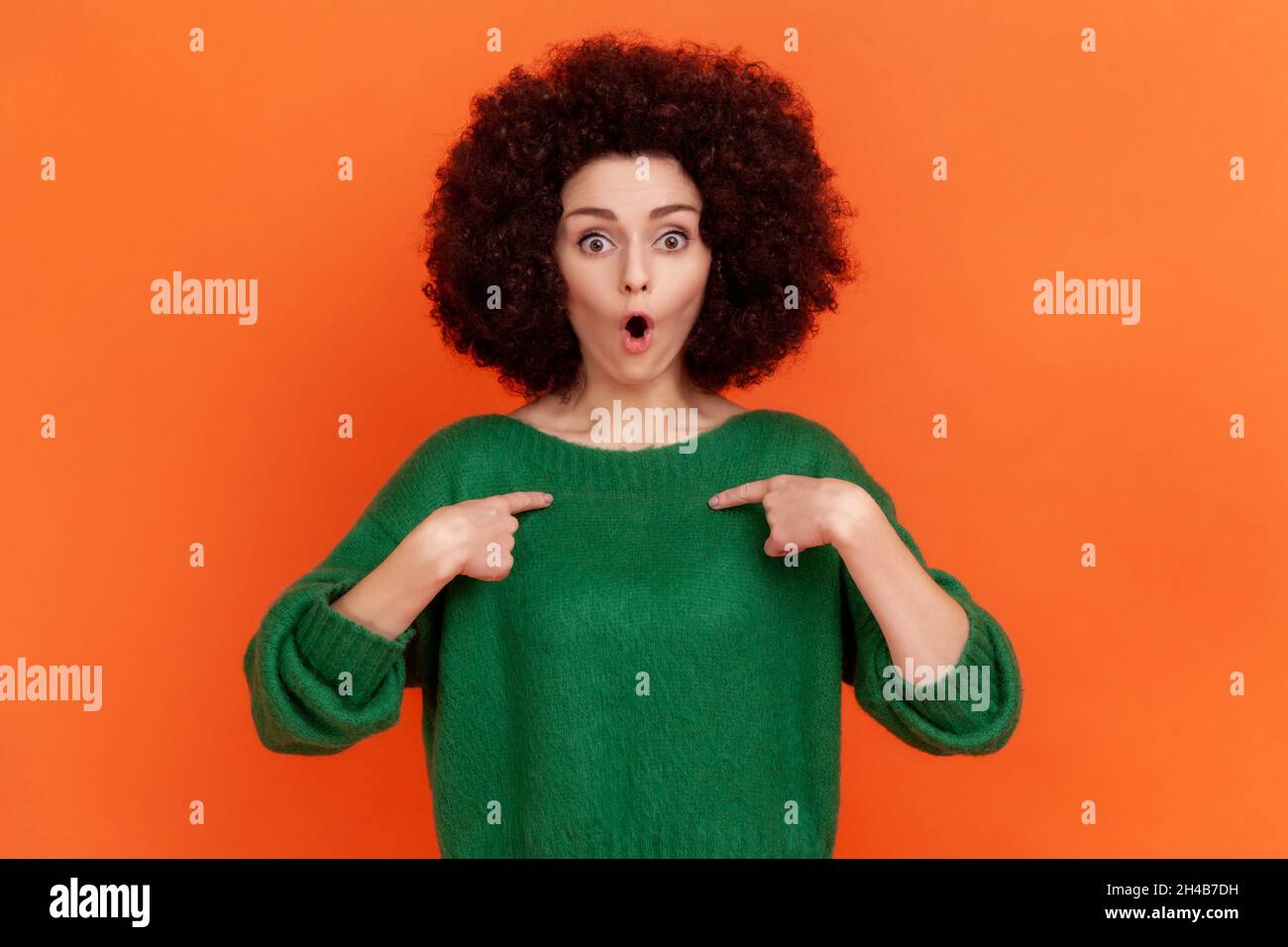 Wow, it is me. Surprised woman with Afro hairstyle in green casual style sweater looking at camera with open mouth, pointing at herself with shock. Indoor studio shot isolated on orange background Stock Photo