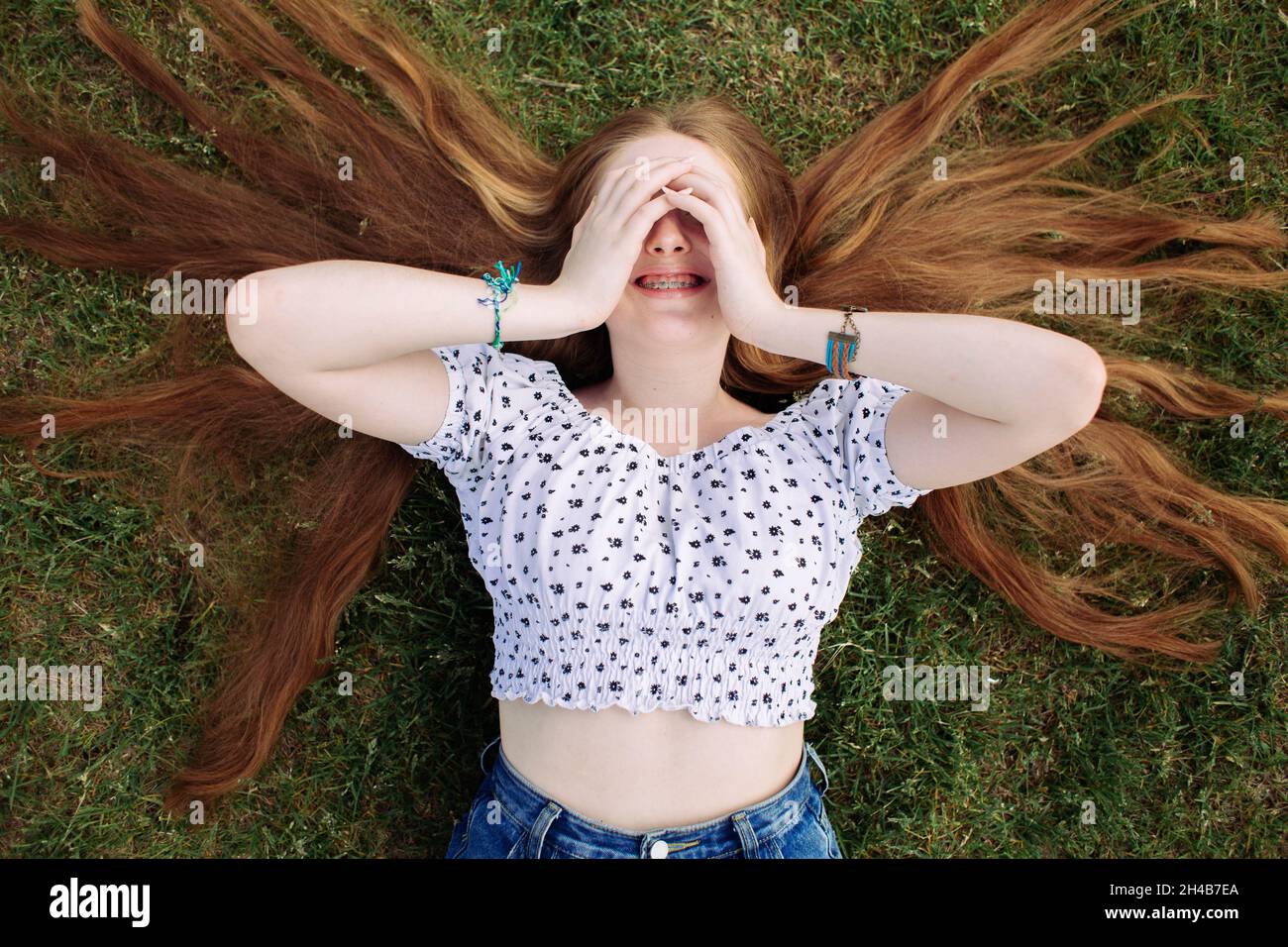Young teen girl with long red hair and braces lying on the green lawn, cover her eyes with hands and laughing Stock Photo