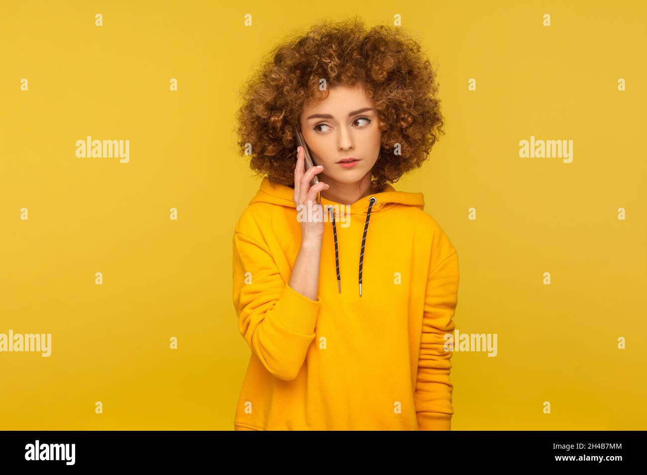 Serious woman with Afro hairstyle using mobile phone, making call and talking with friend, connection, wearing casual style hoodie. Indoor studio shot isolated on yellow background. Stock Photo