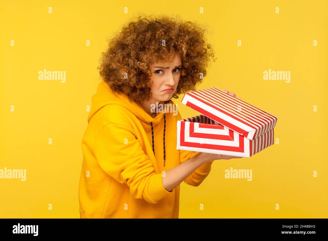 Displeased unhappy woman with curly hair unpacking gift box and looking at camera with frustrated dissatisfied expression, wearing casual style hoodie. Indoor studio shot isolated on yellow background Stock Photo