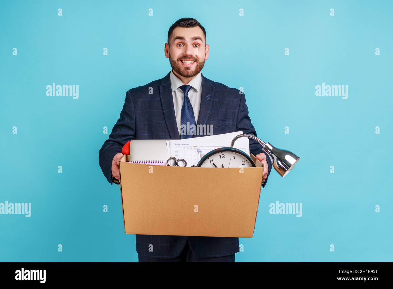 Extremely happy young adult man with beard wearing dark official style suit, stands smiling holding big cardboard box with his stuff, getting new job. Indoor studio shot isolated on blue background. Stock Photo