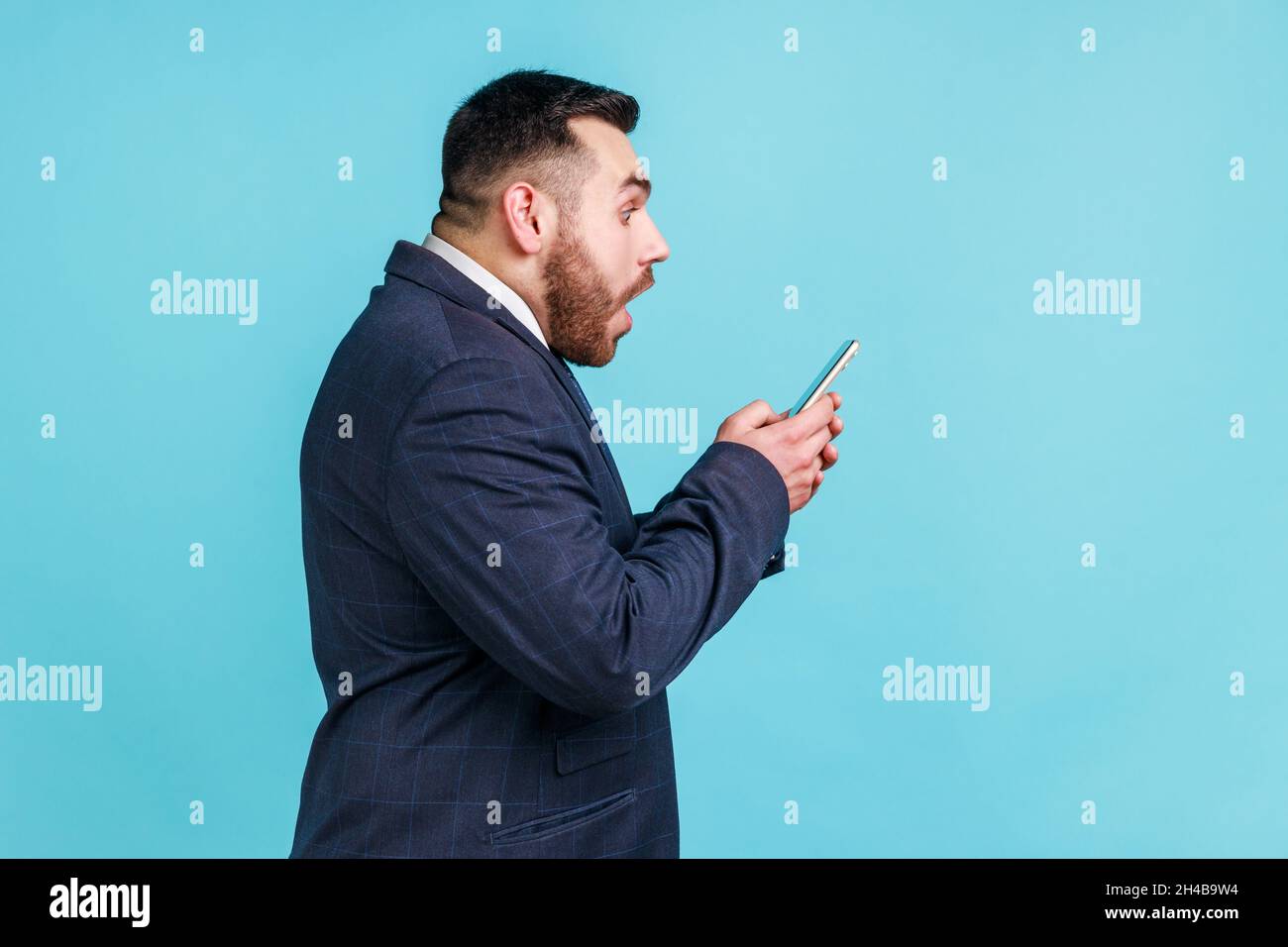 Side view of amazed businessman wearing dark official style suit using online service on mobile phone and expressing amazement, shock. Indoor studio shot isolated on blue background. Stock Photo