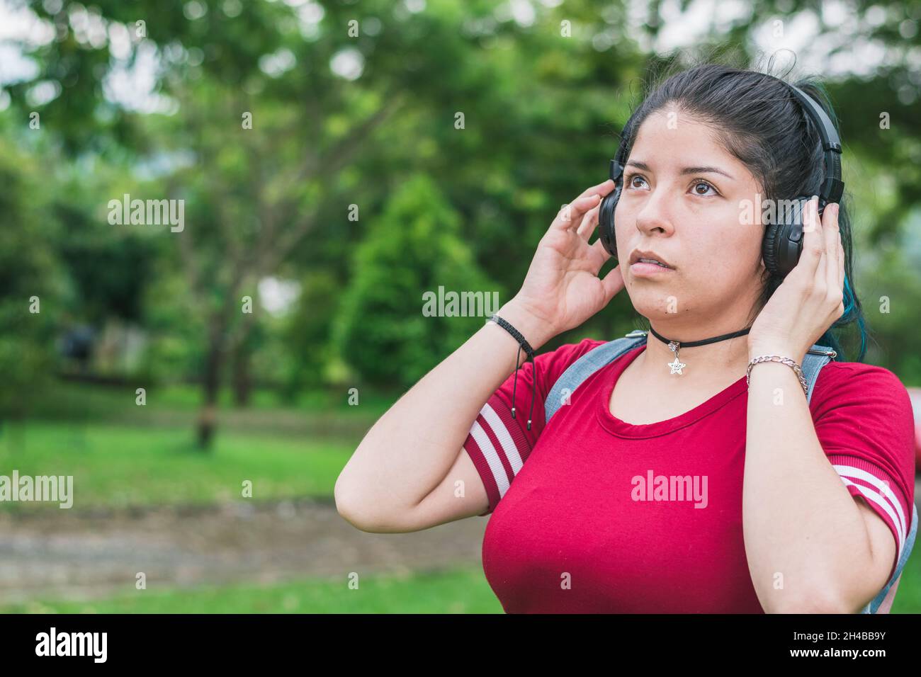 beautiful college woman with blue hair and red dress, listening to music with her wireless headphones in a park, singing passionately looking up with Stock Photo