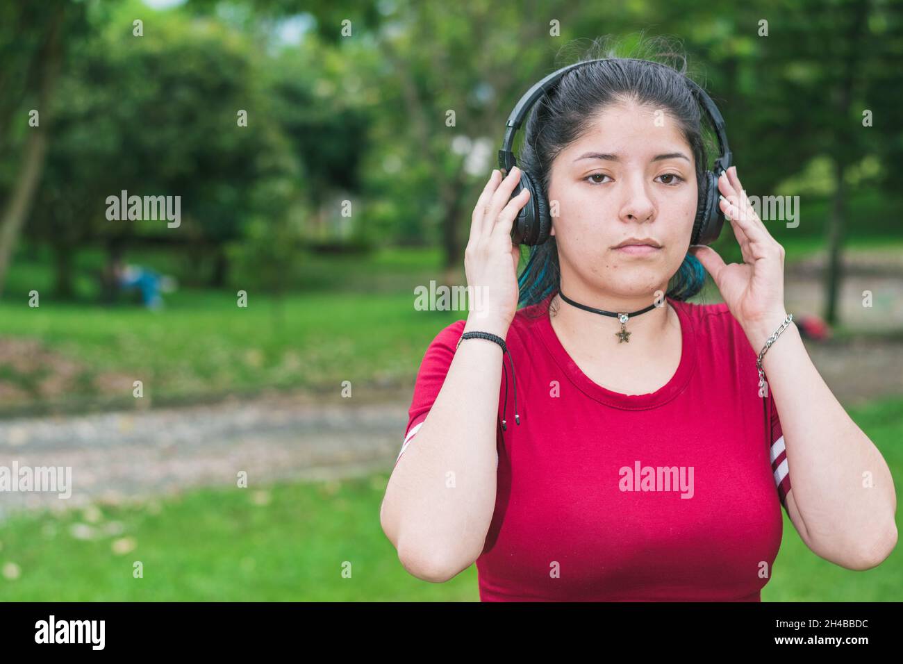 beautiful blue-haired college woman in a red dress, listening to music on her wireless headphones in a park, concentrating, looking straight ahead wit Stock Photo