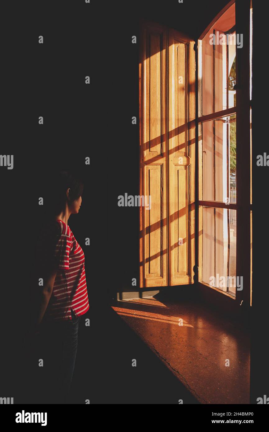 Asian woman standing alone in a dark room looking out of the wooden frame glass window. Dark moody atmosphere. Stock Photo