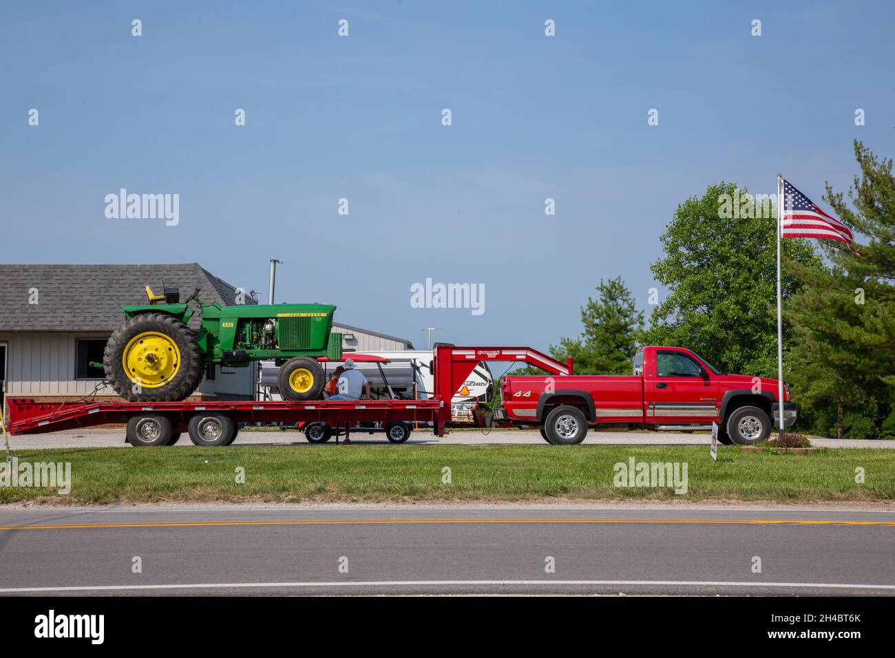 A green John Deere 4520 row crop farm tractor sits on a red flatbed trailer being pulled by a red Chevrolet 2500HD pickup truck in Warren, Ind., USA. Stock Photo