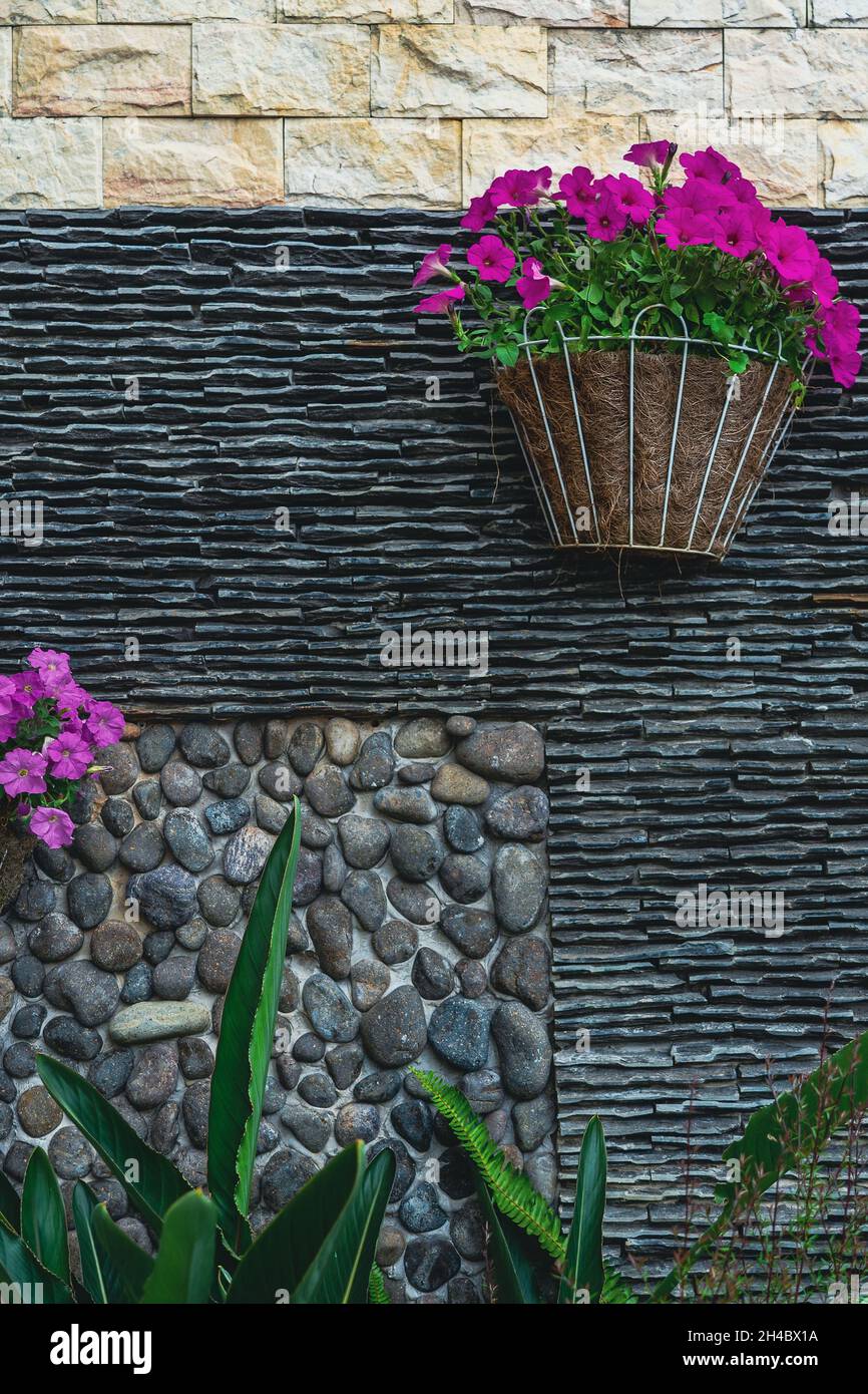 Wall is decorated with grey stones and tiles, and hanging flower pots. Outdoor garden decoration.  Summer garden decoration on backyard. Stock Photo