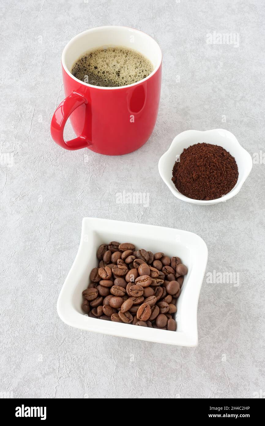 Whole and ground coffee beans in white bowls and black coffee in a red mug on gray concrete. High angle view. Stock Photo