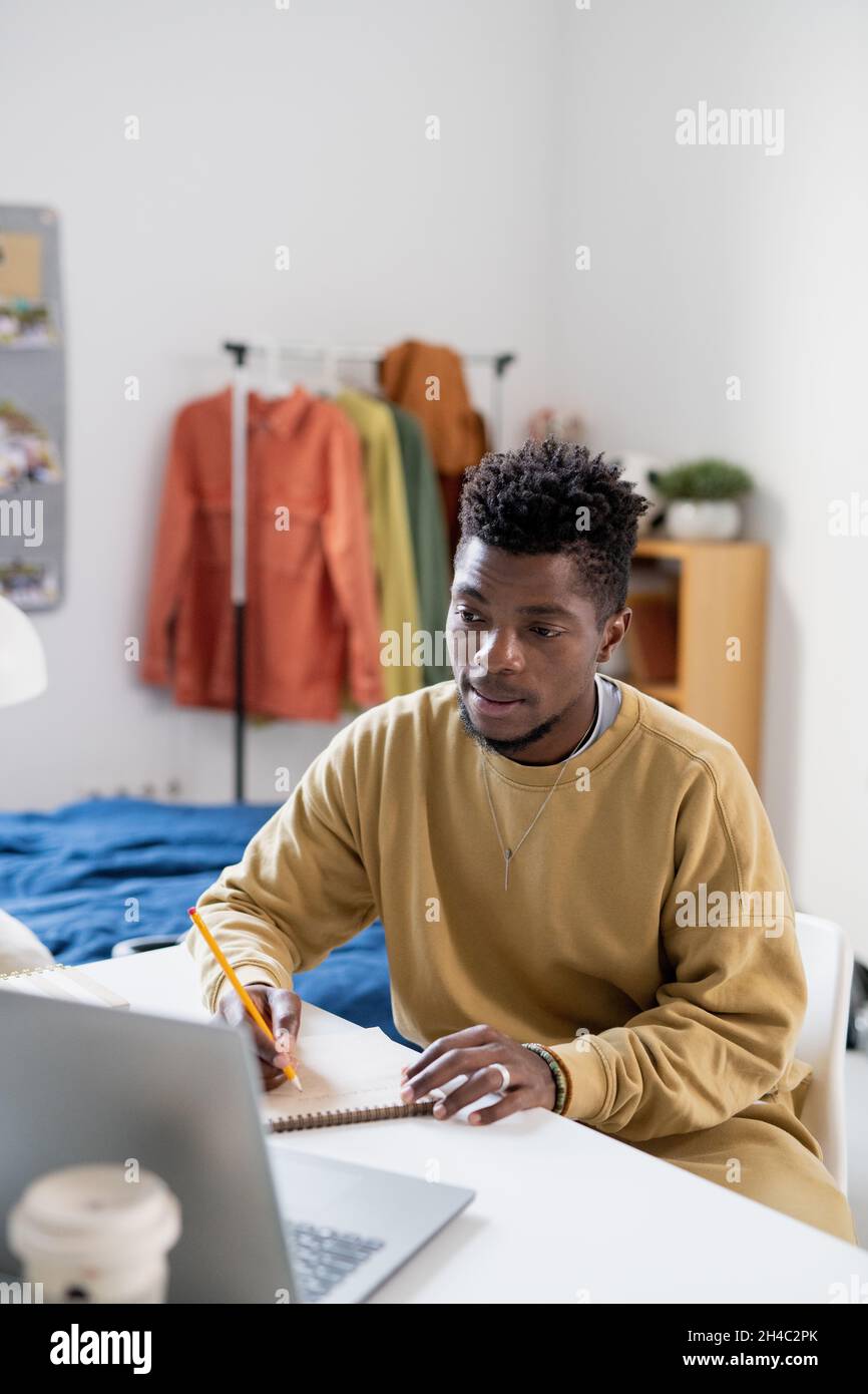 African guy in casualwear looking at laptop display while making notes during online lesson at home Stock Photo