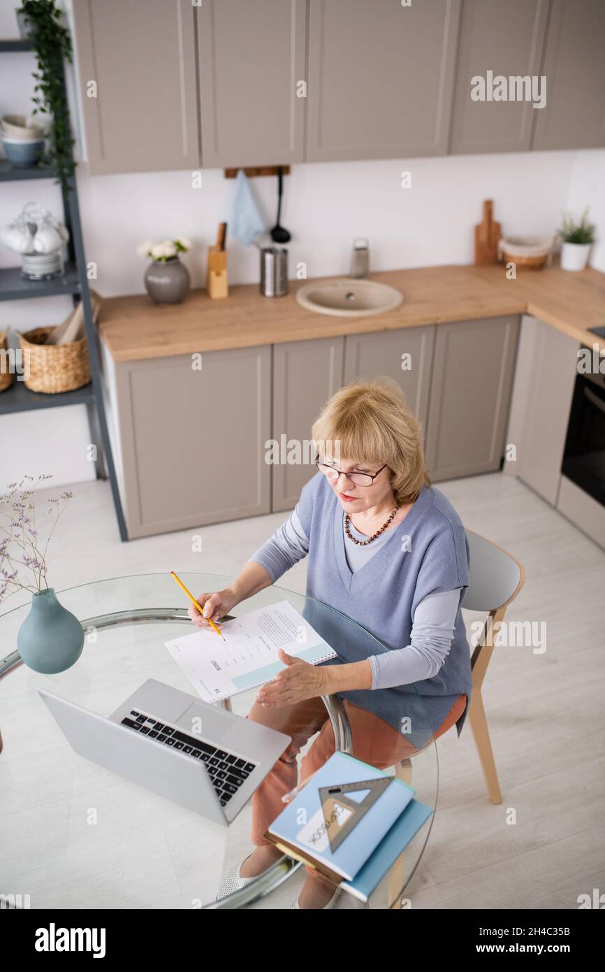 Mature female student communicating with teacher during online course in front of laptop while sitting by kitchen table Stock Photo