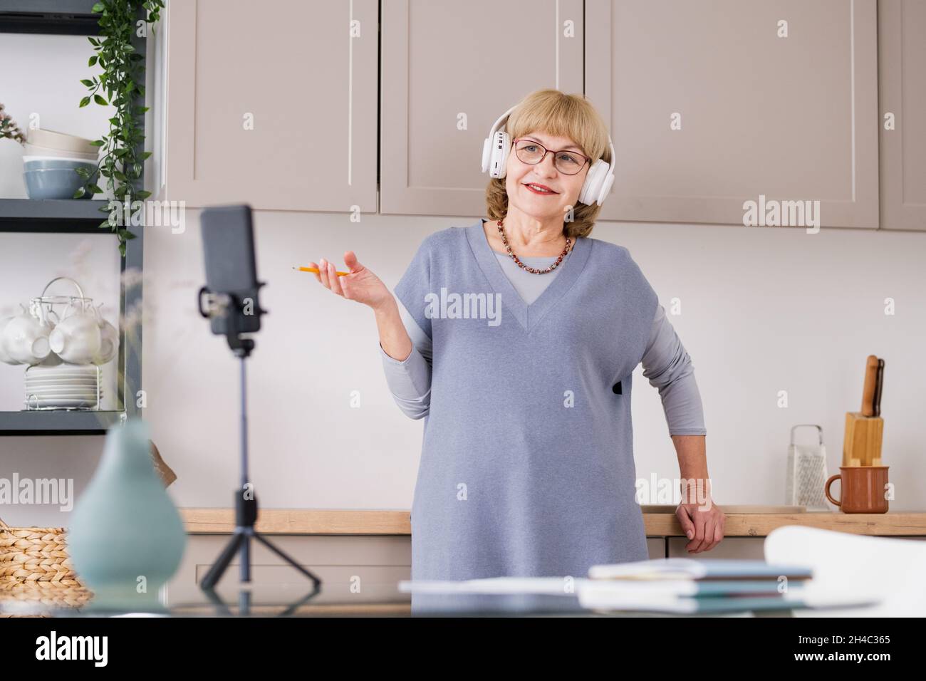 Happy mature female student in headphones and casualwear talking in front of smartphone camera in the kitchen Stock Photo