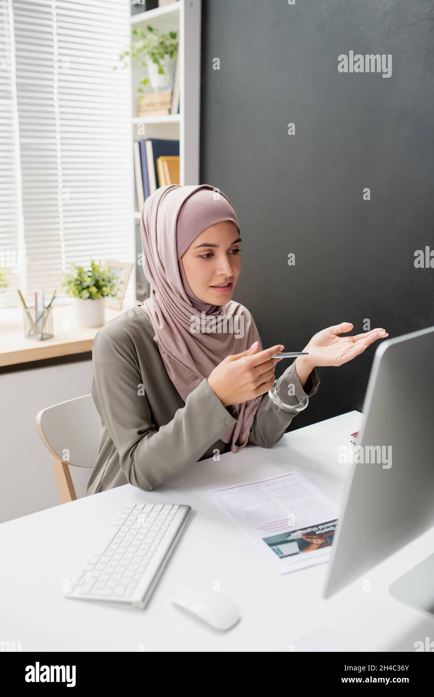 Young confident teacher in hijab speaking to online audience in front ...