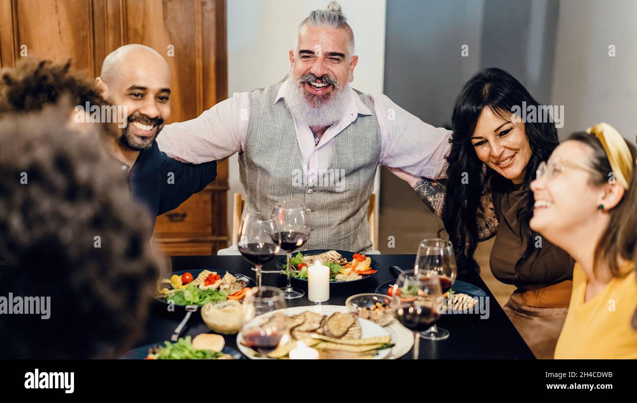 Adult cheerful friends hug happily, sitting at the table during a dinner. Selective focus on the bearded man in the center - high iso image. Stock Photo
