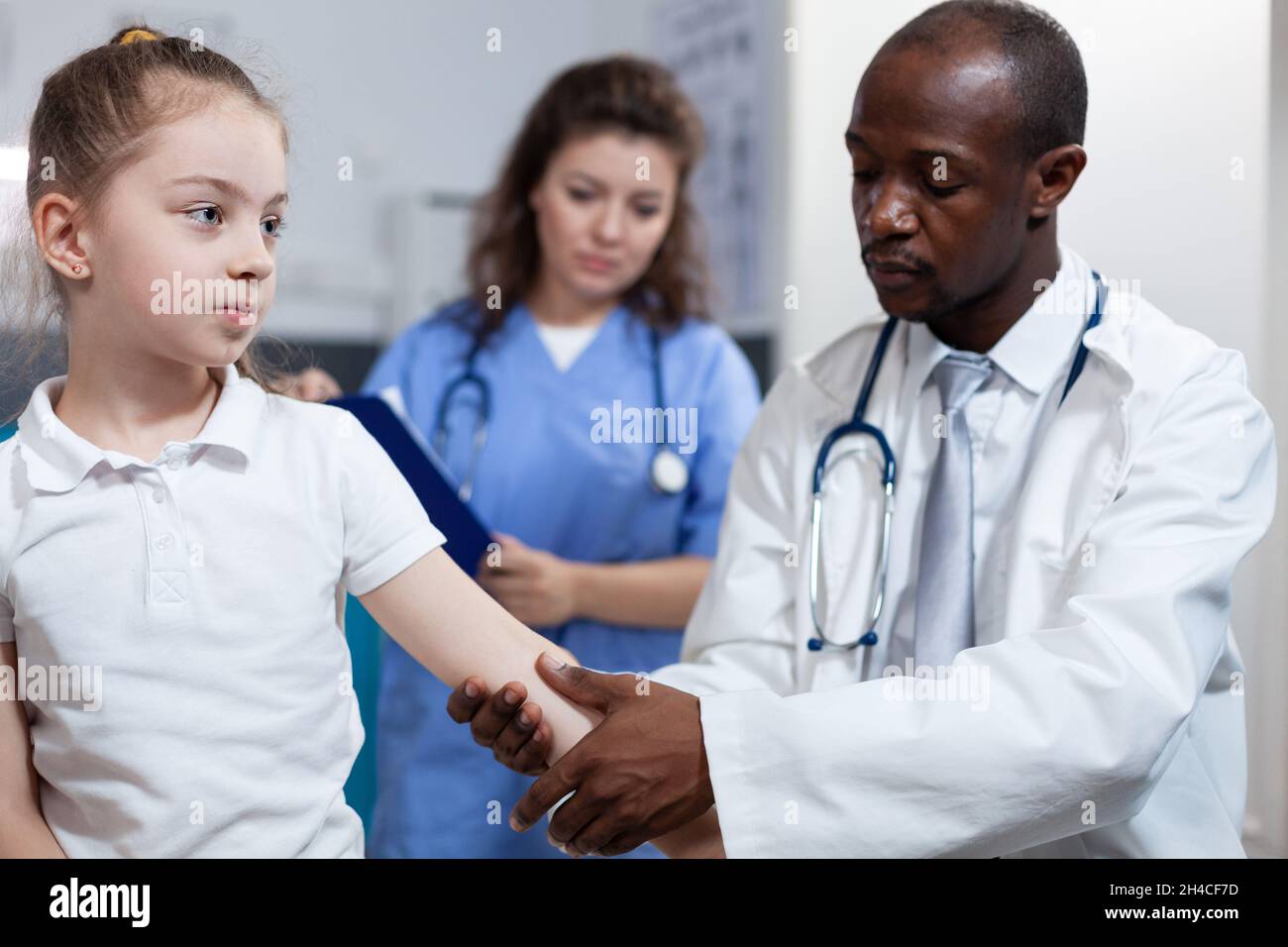 African american pediatrician doctor rubbing fractured arm of little girl during medical physiotherapy in hospital office. Young child having broken bone after accident. Health care service Stock Photo