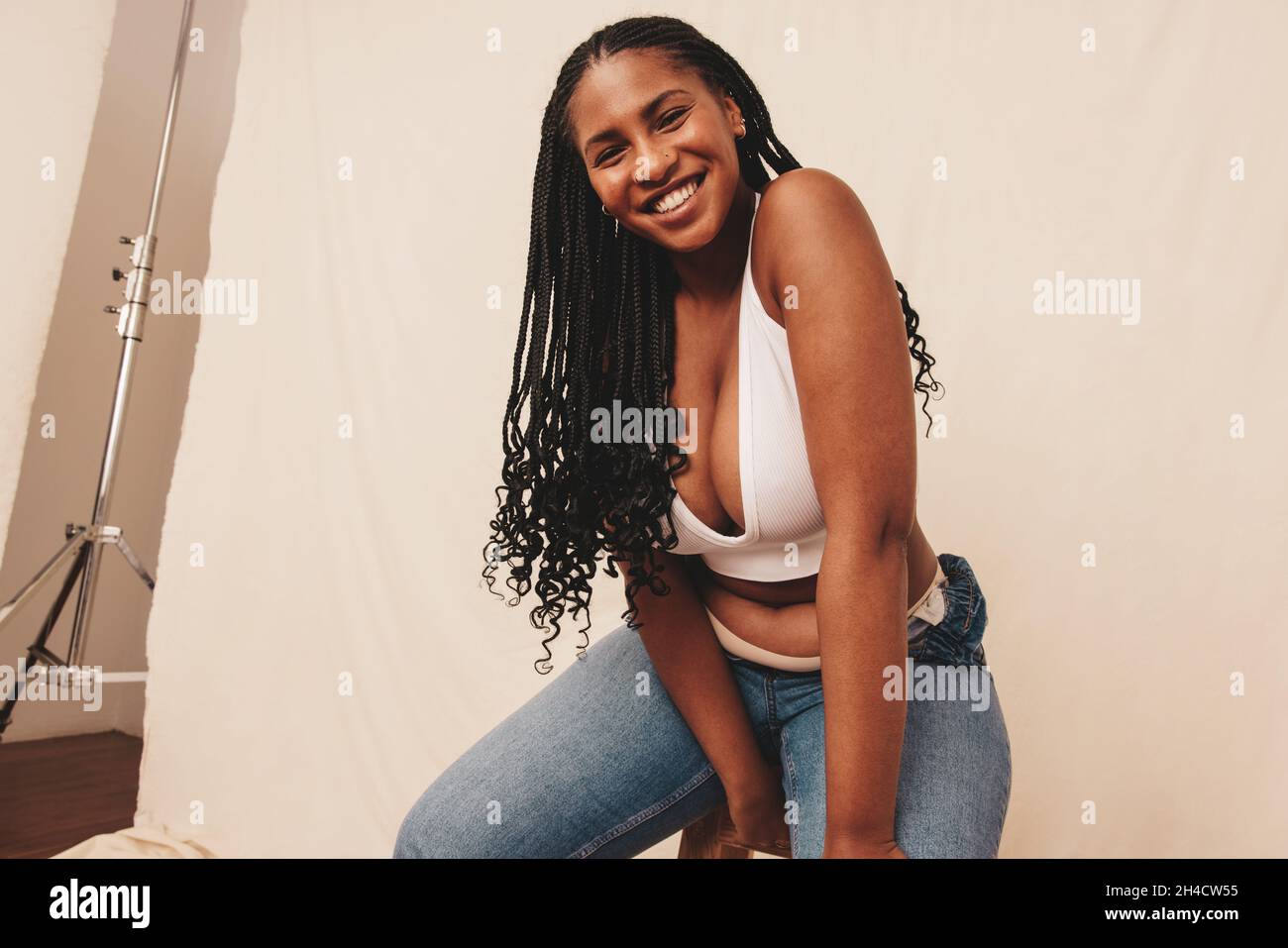 Smiling woman looking at the camera while sitting on a chair in a studio. Happy young woman feeling comfortable in denim jeans and a bra. Body positiv Stock Photo