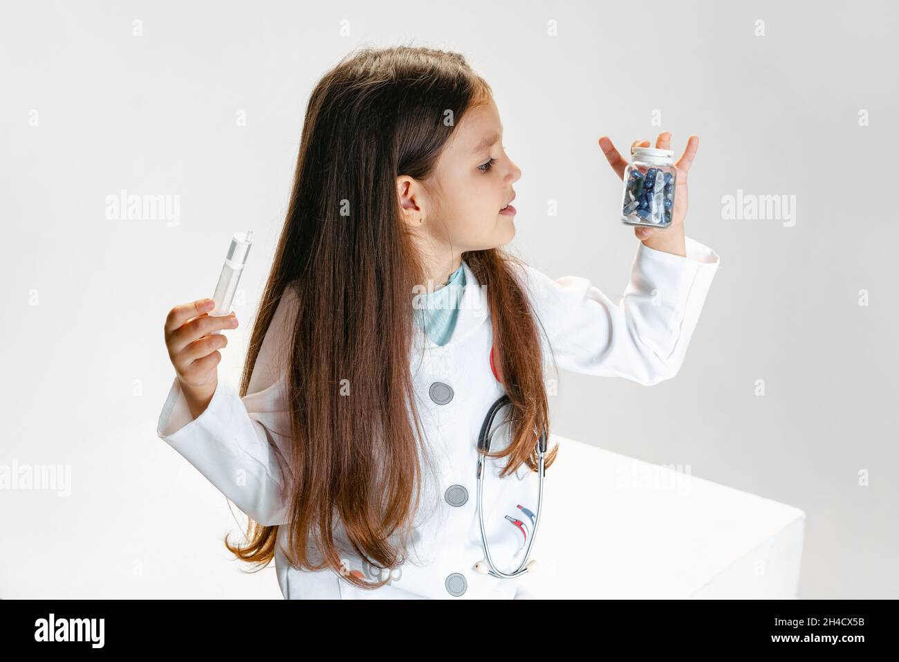 Flyer with cute little girl, child in image of doctor wearing white medicine coat holding pills isolated on white studio background Stock Photo
