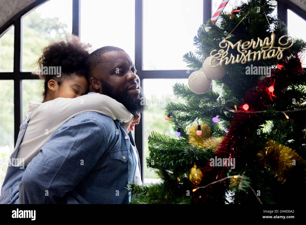 Beautiful young african american girl being picked by handsome father decorating christmas tree during festival by adding candy cane at the top of the tree near a star at home Stock Photo