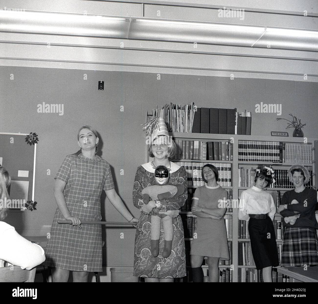 1960s, historical, some female students at a business college in a corner of a library standing by the bookcases filled with encyclopedias and reference books, having fun roleplaying with soft toys, Monroe College, Middletown, Virginia, USA. One of the girls is holding a batman soft toy. A sign on top of one of the bookcases says THINK. Stock Photo