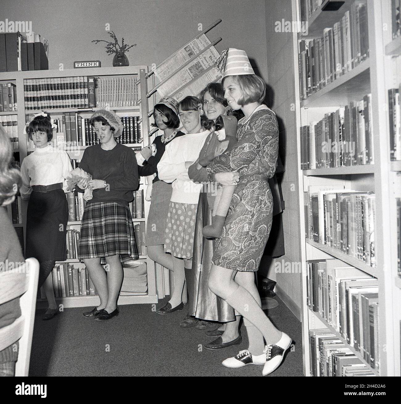 1960s, historical, some female students at a business college in a corner of a library standing by the bookcases filled with encyclopedias and reference books, having fun roleplaying with soft toys, Monroe College, Middletown, Virginia, USA. One of the girls is holding a batman soft toy. A sign on top of one of the bookcases says THINK. Stock Photo