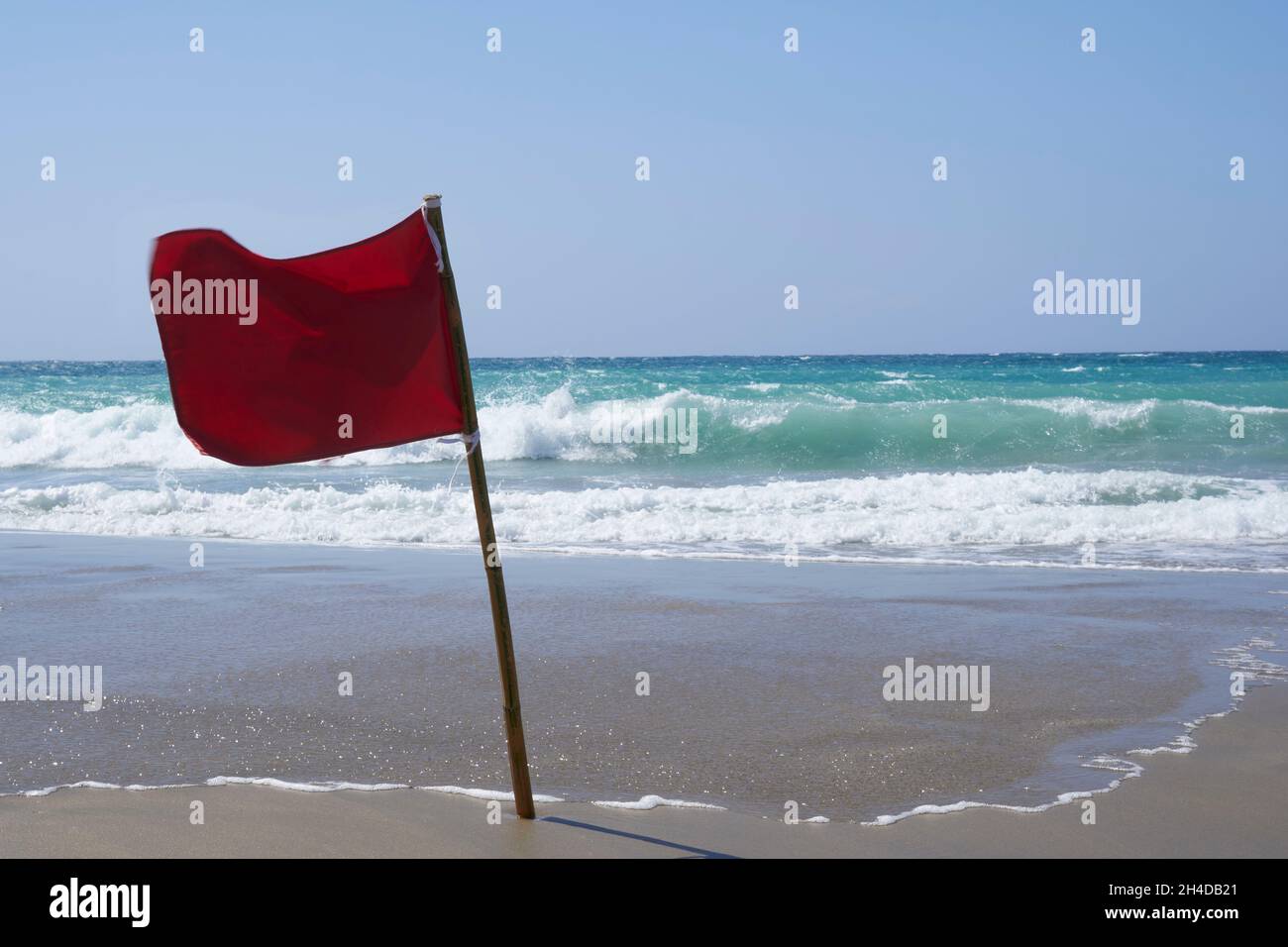 Red warning flag blowing in wind on beach with rough surf in background Stock Photo