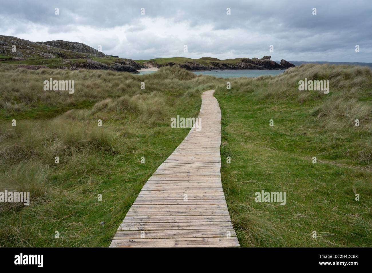 Clachtoll beach in Assynt, Scottish Highlands on a cloudy day with dramatic sky. Wooden boardwalk leading through grass to beach. Stock Photo