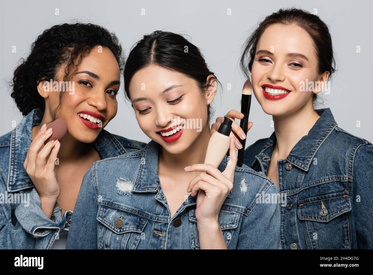 Smiling interracial women holding face foundation, cosmetic brush and beauty blender isolated on grey Stock Photo