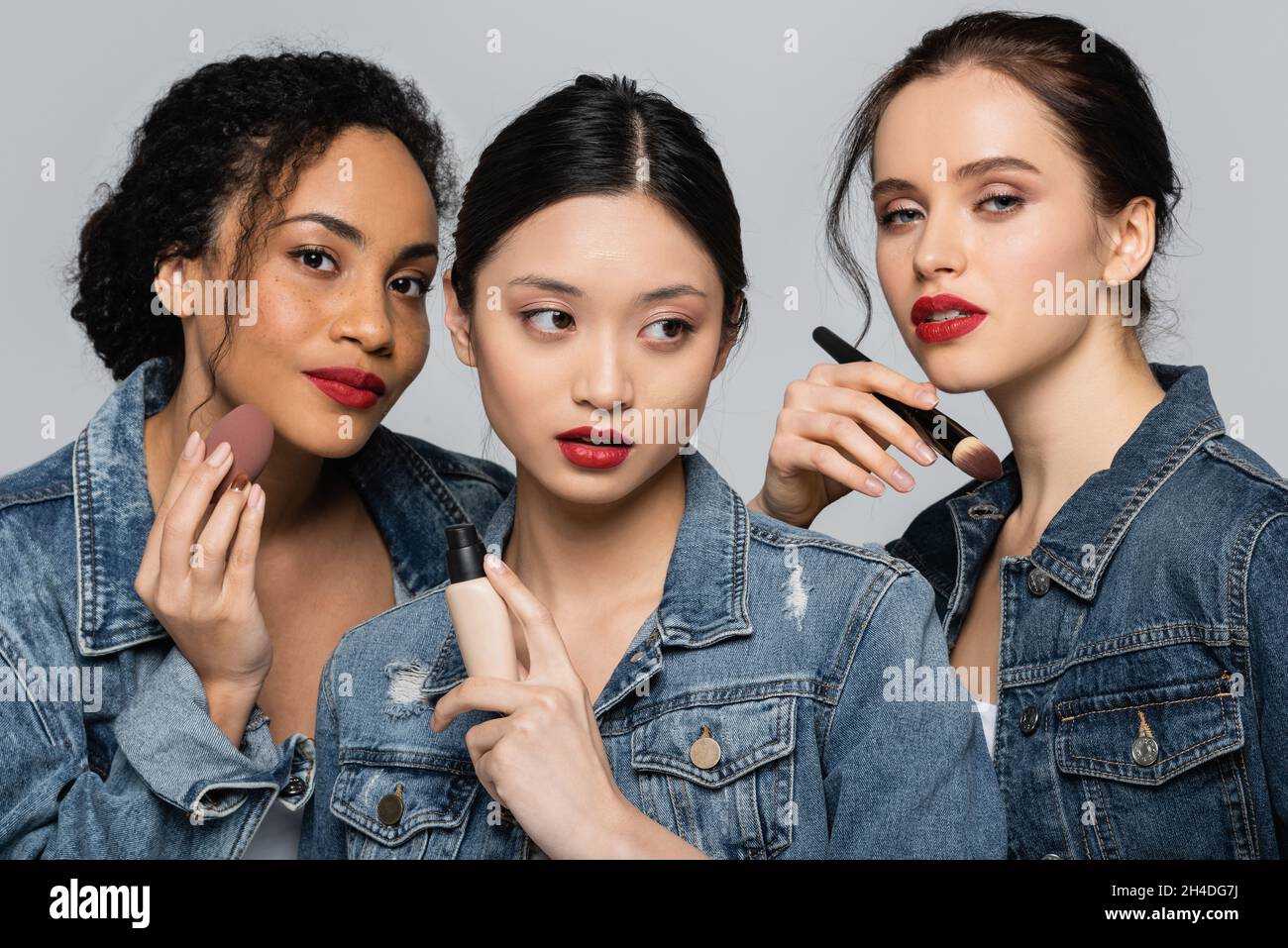 Interracial young women holding cosmetic brush, face foundation and beauty blender isolated on grey Stock Photo