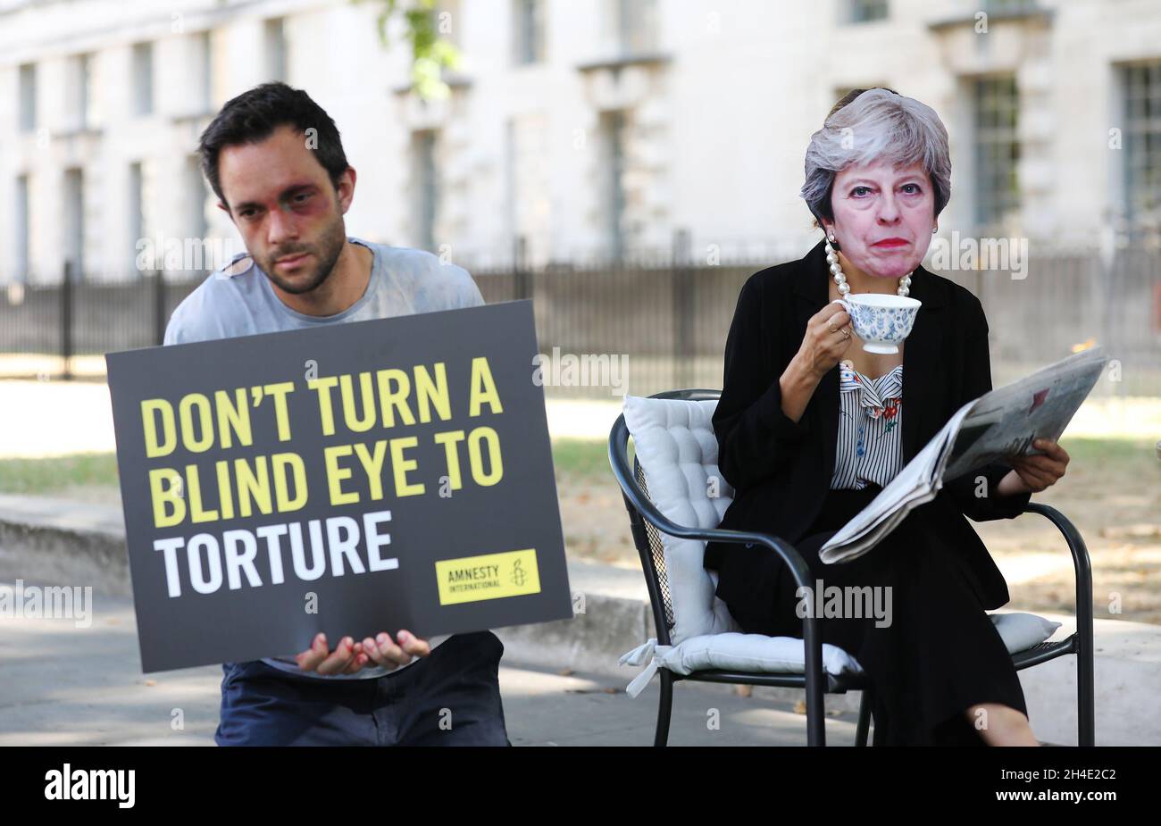 An activist wearing a mask of Prime Minister Theresa May stages a photocall outside Downing Street as she drinks a cup of tea and reads the paper while ignoring three torture 'victims' in stunt which highlights a call by Amnesty International for inquiry into UK's involvement in torture. Picture dated: Monday August 6, 2018 Stock Photo