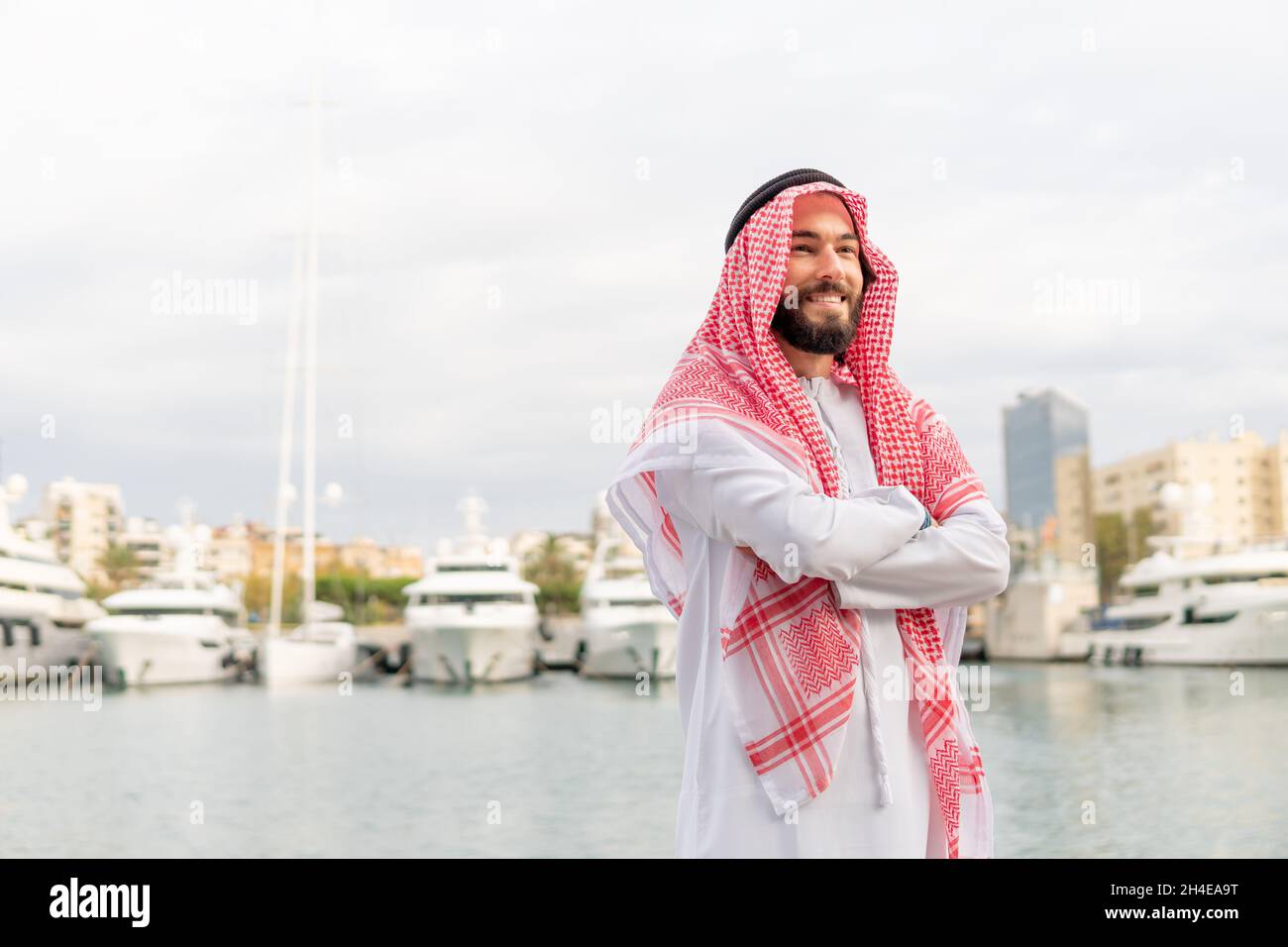 Happy Arab man in keffiyeh crossing arms and looking away with smile while standing near water on sunny day in city port Stock Photo