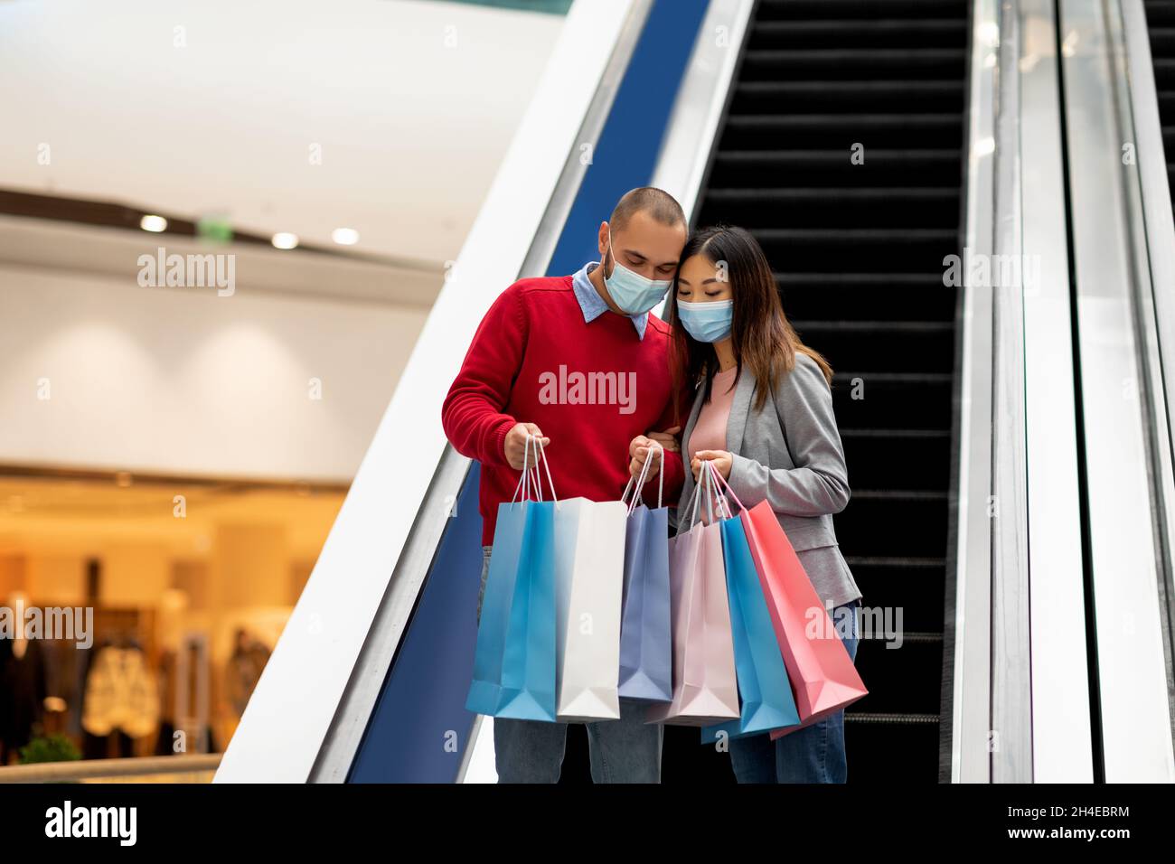 Young international couple in protective masks going down on escalator at mall, looking inside shopping bags, free space Stock Photo