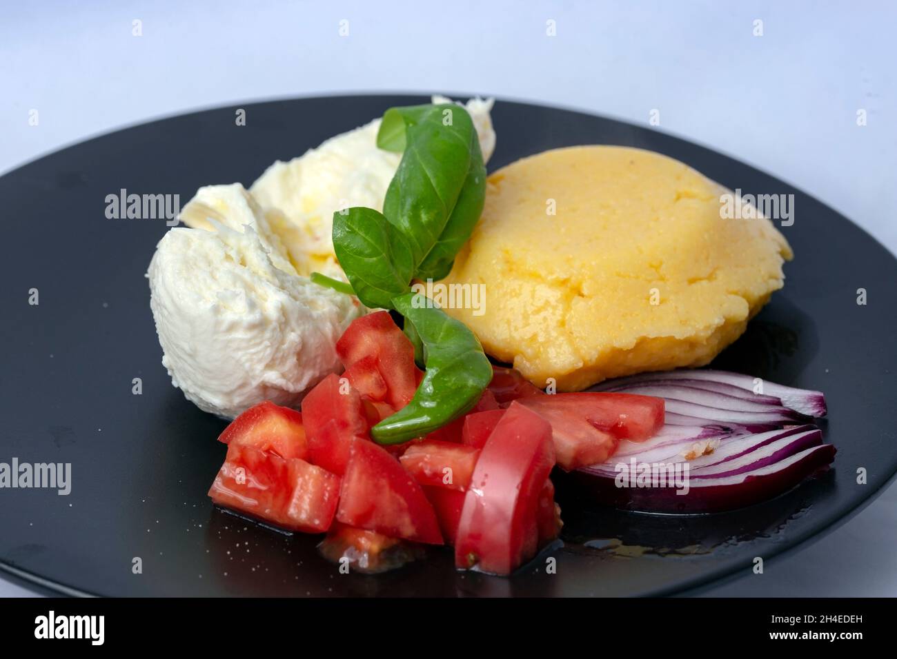 Polenta with mozzarella, sliced tomatoes, basil leaves and red onion, on a black ceramic plate. Side view close up Stock Photo