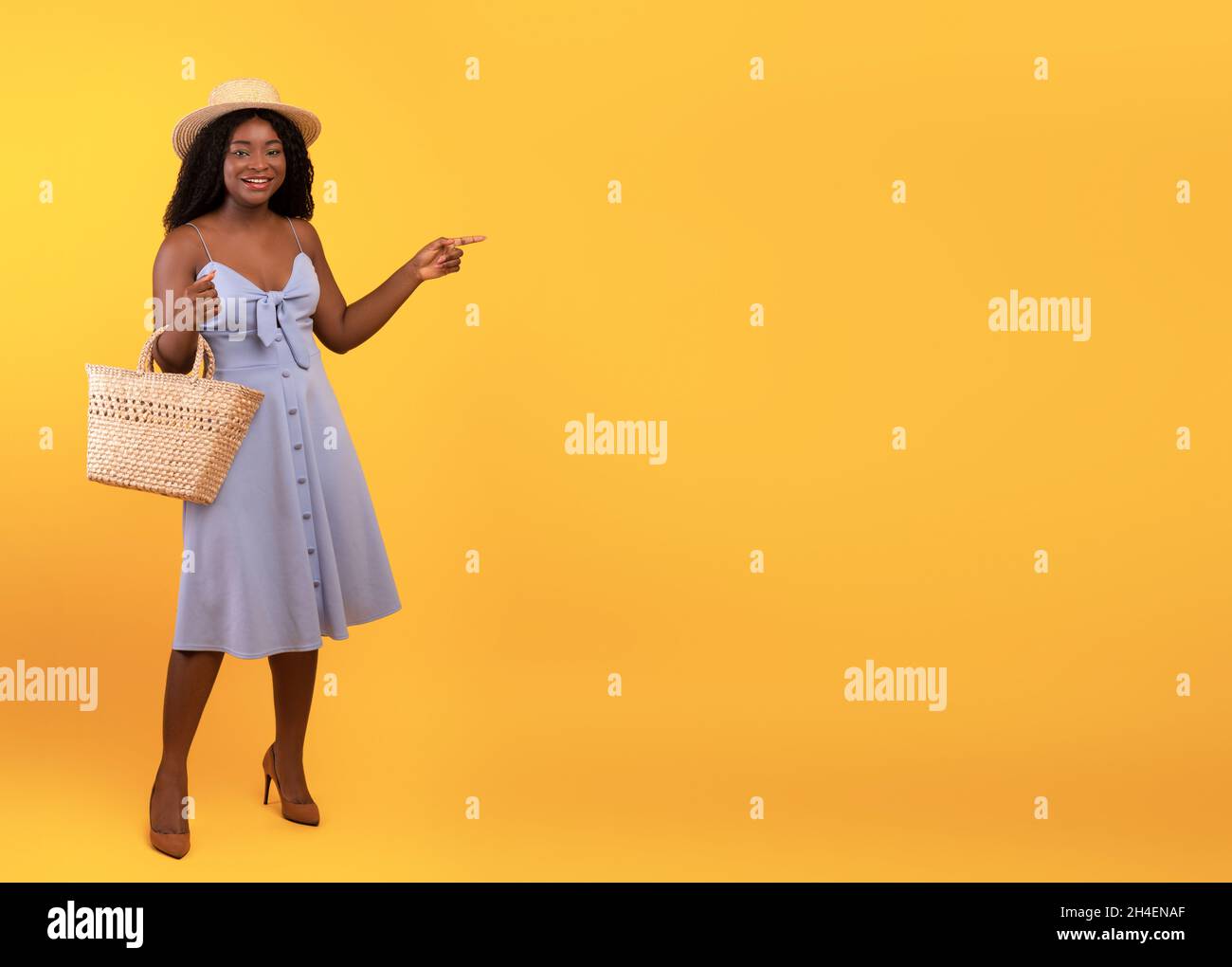 Seasonal shopping. Charming black woman wearing dress and hat, holding straw bag, pointing aside at copy space Stock Photo