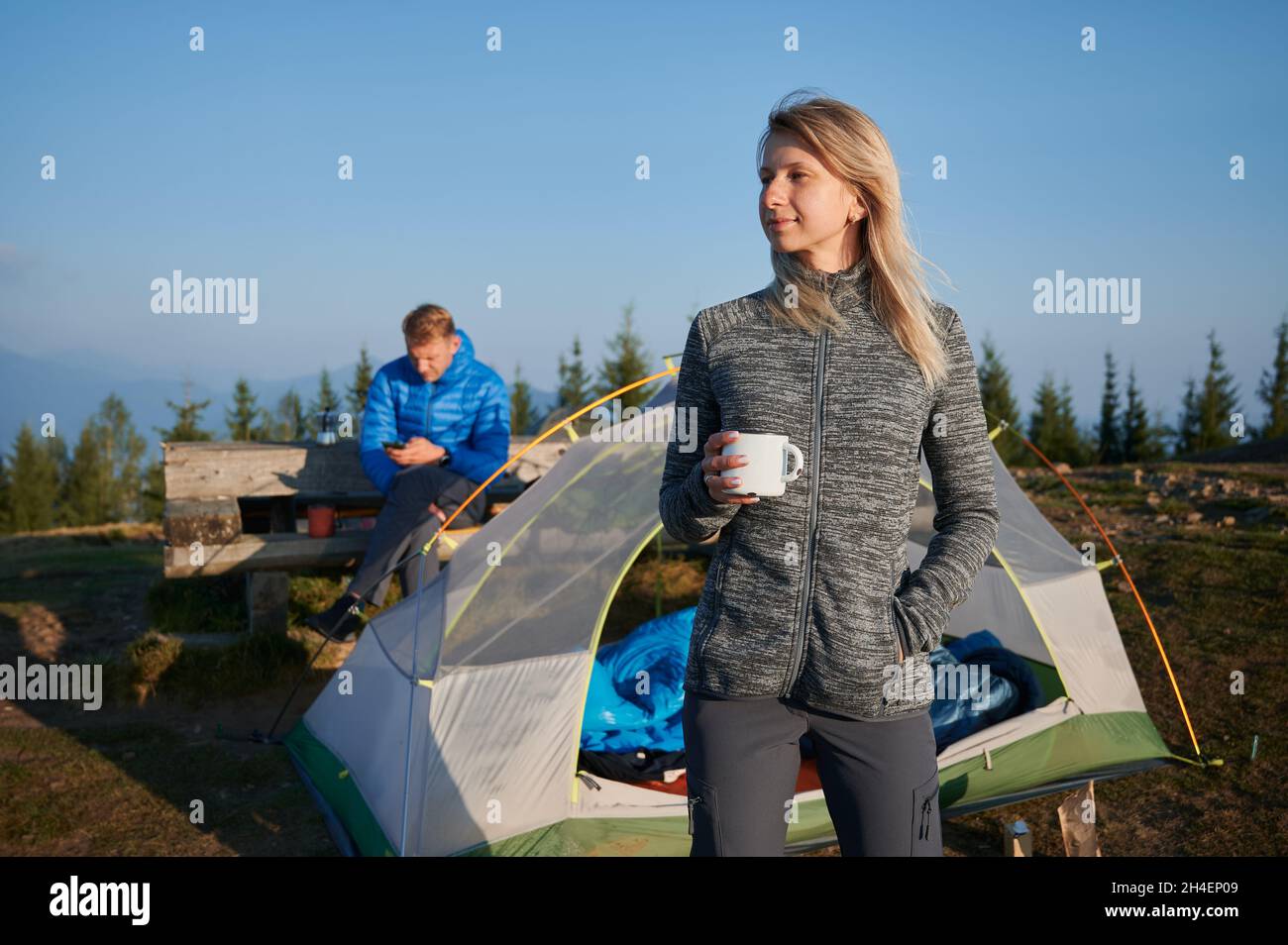 Charming female hiker holding cup of coffee and smiling while man sitting on bench and using smartphone. Young couple travelers resting outdoors neat tourist tent with blue sky on background. Stock Photo