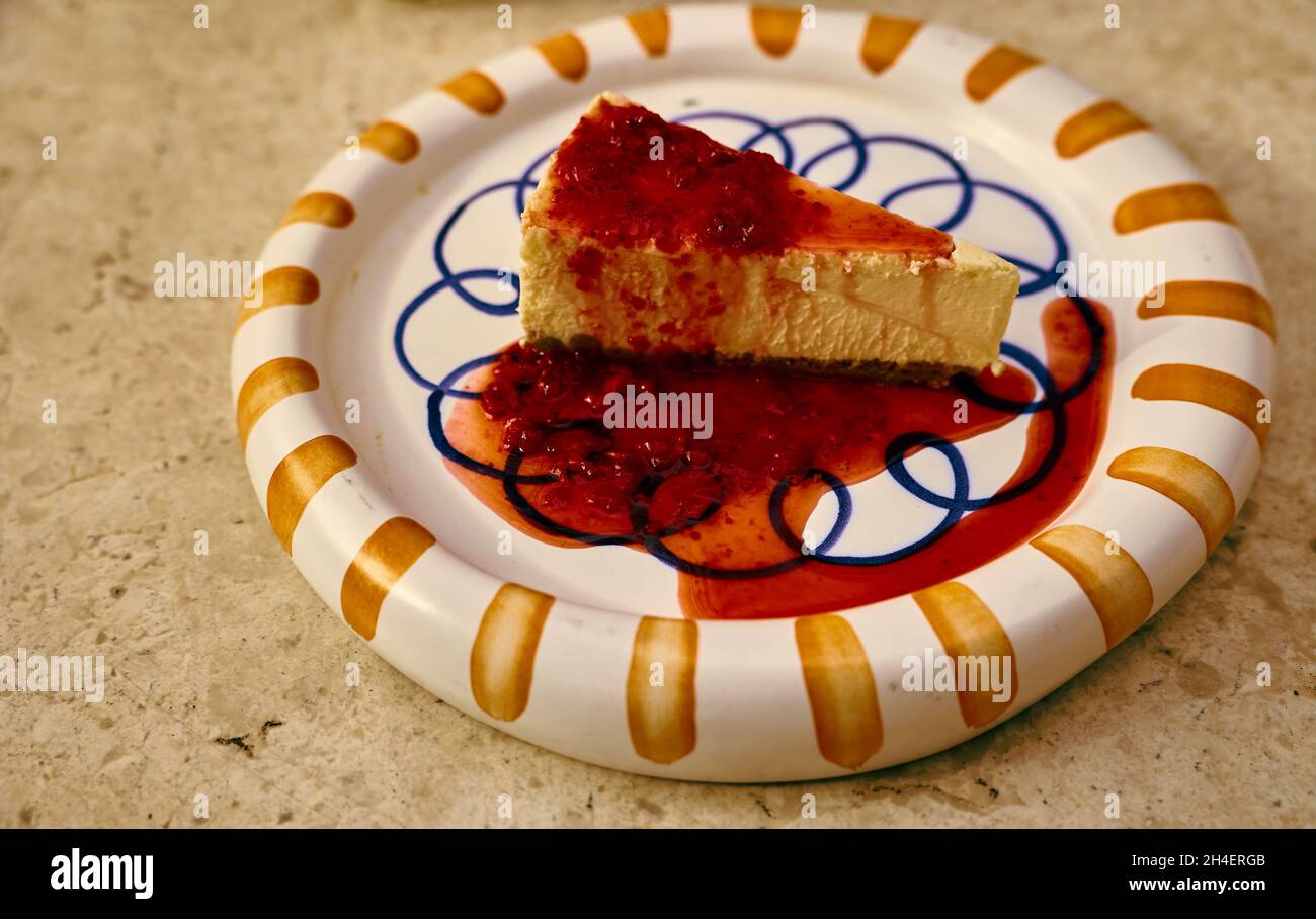 Delicious cheesecake drizzled with raspberry syrup on a painted plate with a restaurant table in the background. Stock Photo