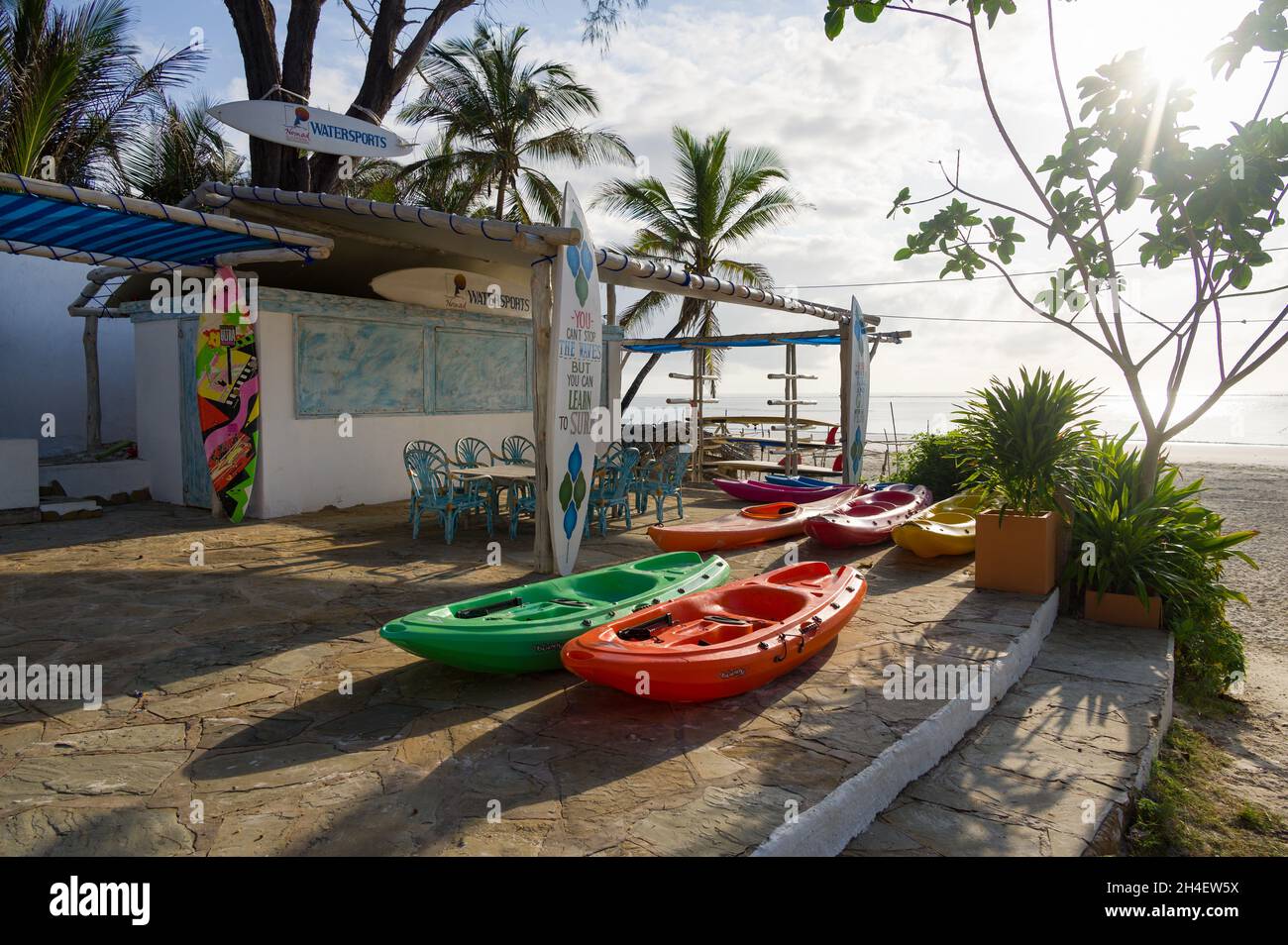 Various watersports equipment on display outside a watersports shop with tropical beach and Indian ocean in background, Diani, Kenya Stock Photo