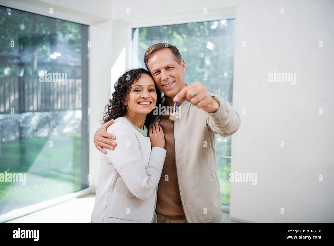 cheerful man holding blurred key and hugging african american wife in ...